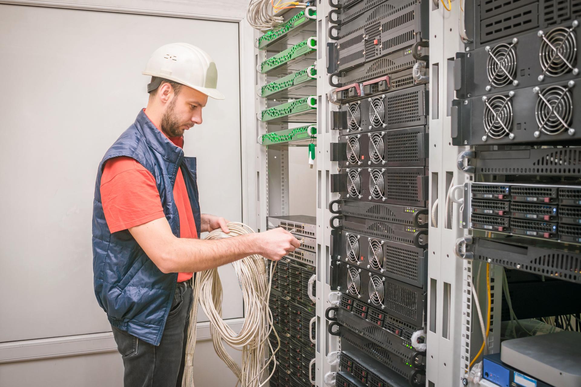 A man works in the server room of a data center. A technician connects a cable to a stack of managed switches. An engineer in a white helmet connects Internet wires to new equipment. Technology concept